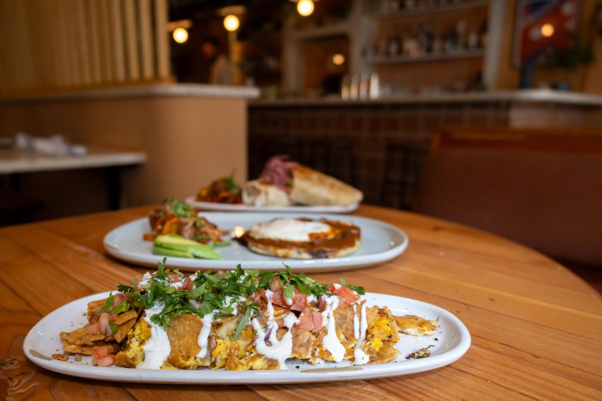 Three white plates on a wooden table: the migas are nearest to the camera, the bean and cheese pupusa is second nearest and the pork shoulder burrito is farthest.