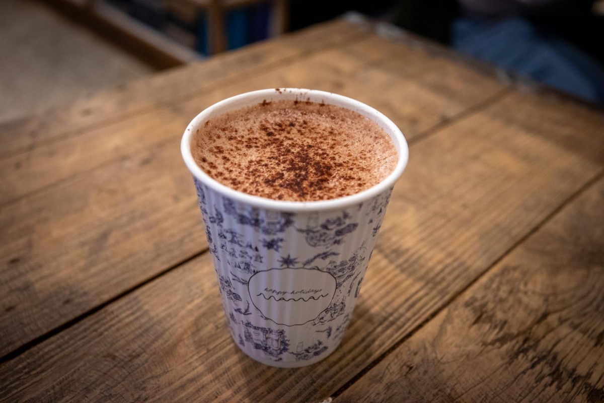 A white-and-blue cup of hot chocolate with cocoa powder in it. It is on a wooden table and on the cup it reads, “happy holidays” and maman.