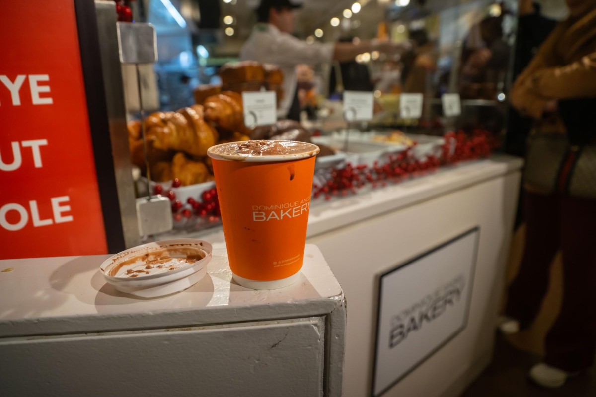 An orange cup of hot chocolate sits next to a white lid on a white ledge. There are pastries in a display case in the background.