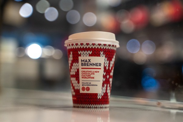 A red-and-white cup of hot chocolate, against a blurry background. The cup has a white lid. On the cup, there is a white box with text that reads, “MAX BRENNER” and “CREATING A NEW CHOCOLATE CULTURE WORLDWIDE.”