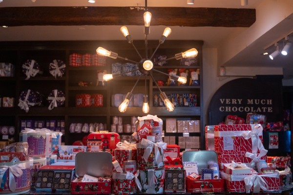 An overhead light with multiple lightbulbs. Underneath, there are boxes of chocolate, some of them wrapped in gift-wrapping paper and tied with white bows. Behind them, there are shelves with more boxes, and to the right there is a sign that reads, “VERY MUCH CHOCOLATE.”