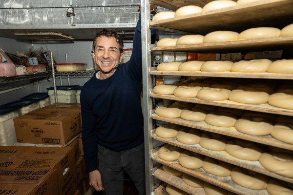 A man in a blue sweater and jeans poses for a photo next to a metal shelf that is full of uncooked bagels.