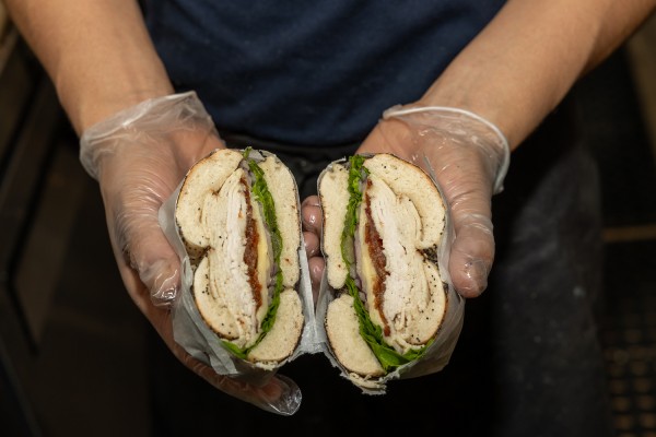 Gloved hands hold a halved bagel sandwich made with sliced meat, lettuce, cheese, onions, and bacon.