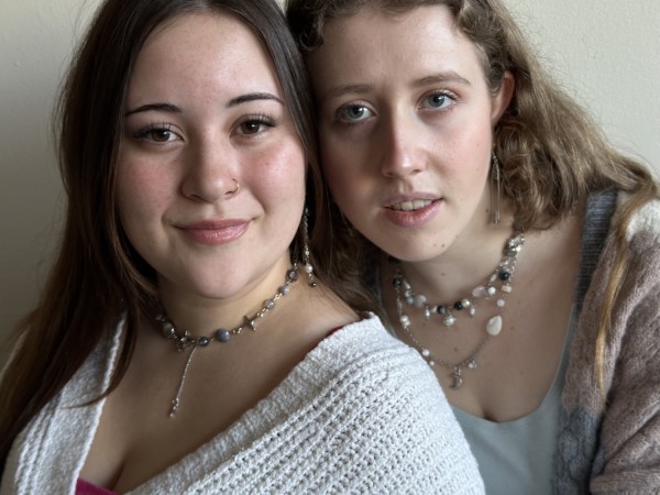 Two women, one wearing a white shirt and the other wearing a gray shirt with a brown-gray sweater standing next to each other in a close-up. Both of them are looking straight at the camera and wearing necklaces with multiple beads and bobbles.