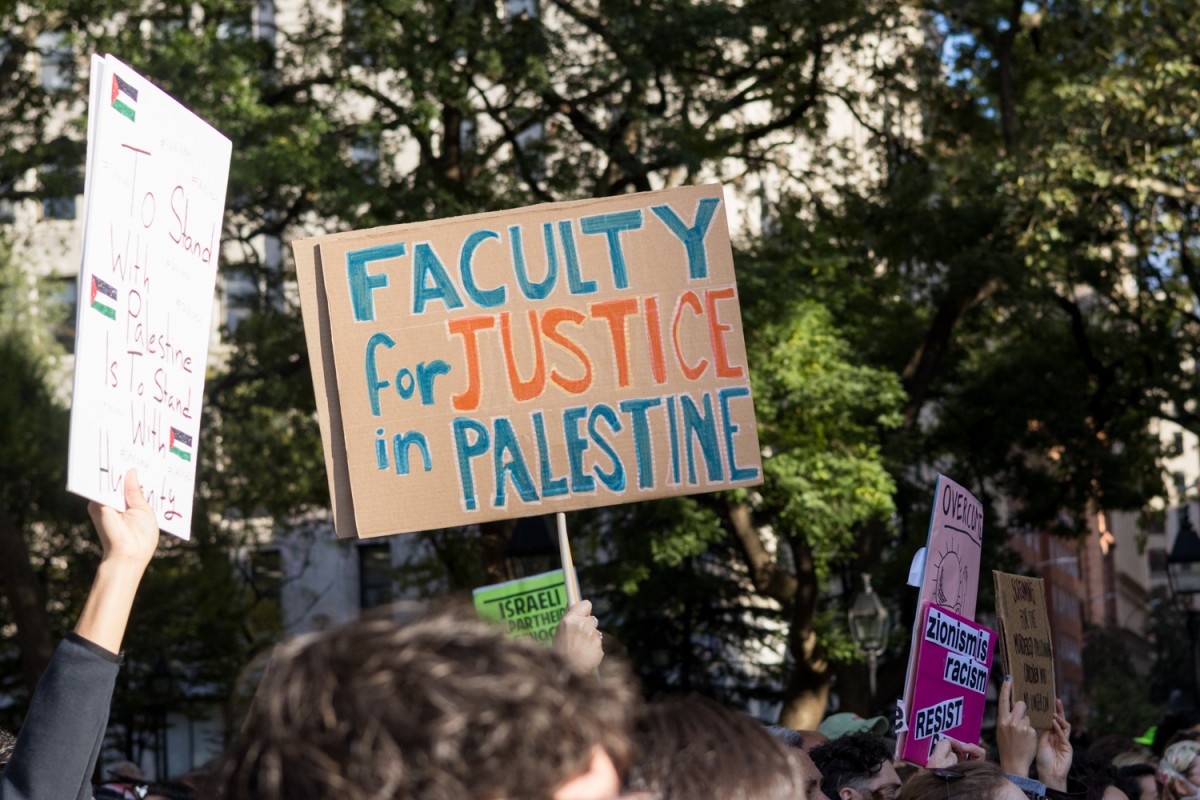 A protester holds a cardboard sign reading Faculty for Justice in Palestine above a crowd in a park.