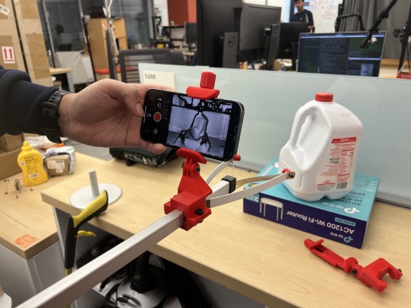 A cell phone displaying an image in black and white of the table in front of it. On the table is a jug of milk. The phone is being held in place by a red clamp and someone’s hand is hitting the record button.