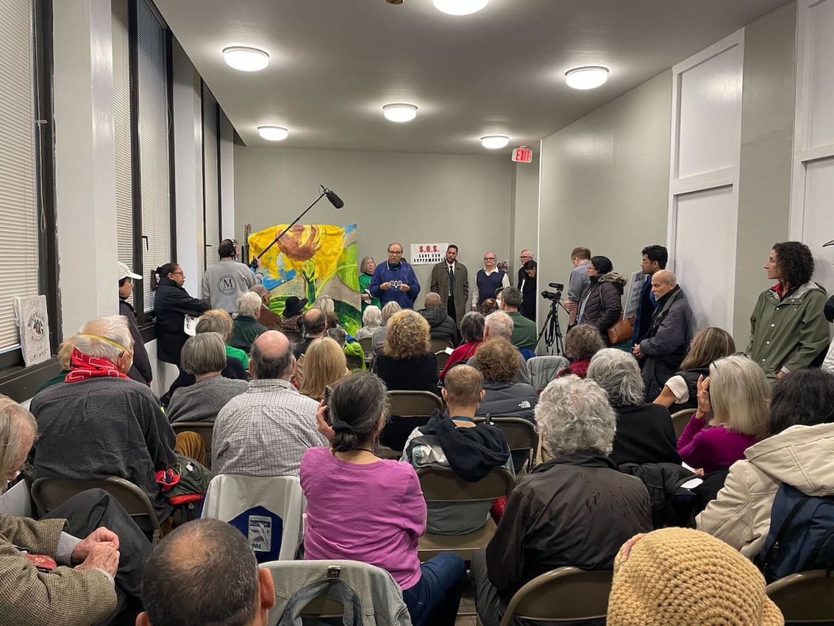 Dozens of people sit in folding chairs facing the front of the room. A man speaks at the front of the room next to a poster of a mural. Multiple reporters are filming them.