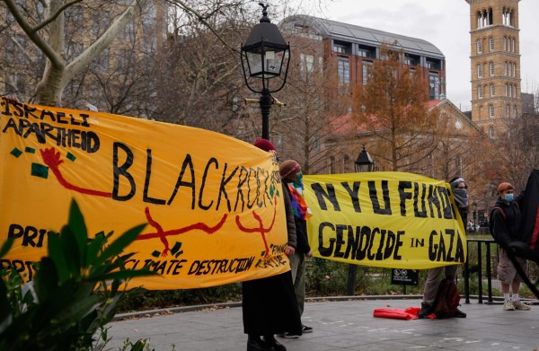 A group of people hold two large banners on a stage in Washington Square Park. On the left is an orange banner with “BLACKROCK” written in the middle, and drawing of red hands pointing to words such as “ISRAELI APARTHEID” and “CLIMATE DESTRUCTION.” On the right is a yellow banner that reads “N.Y.U. FUNDS GENOCIDE IN GAZA”.