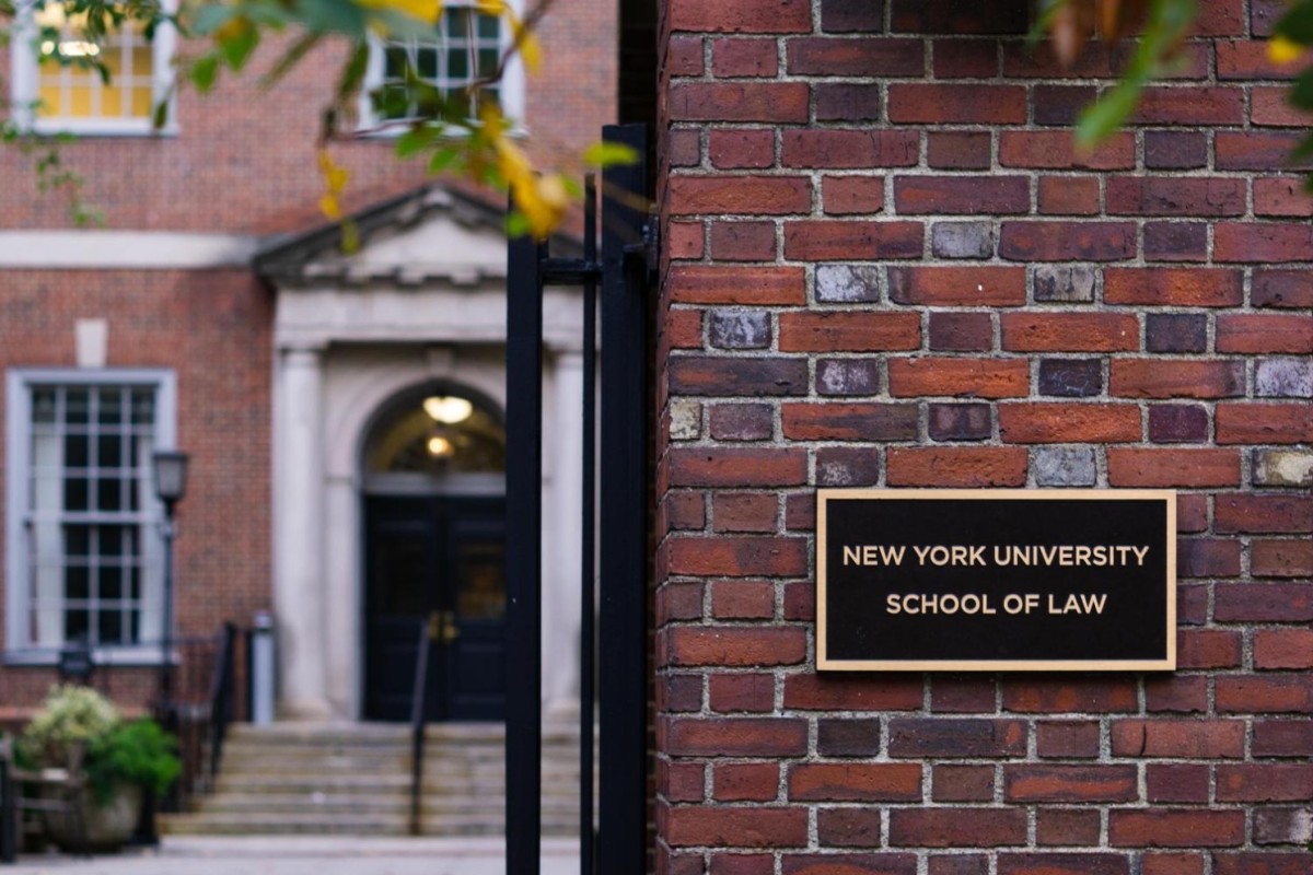 To the right there is a sign on a brick wall that reads, ‘New York University School of Law.’ Behind that wall is the entrance to a brick building.