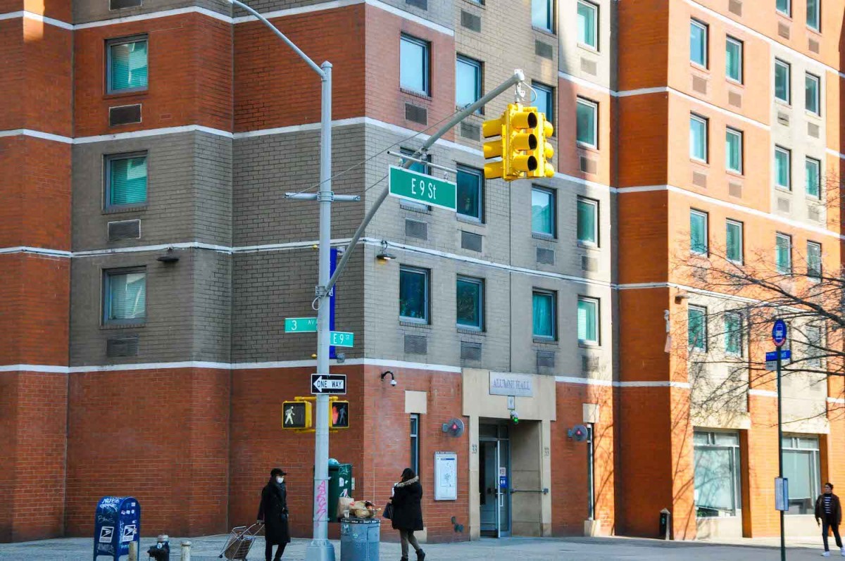 A building facade with gray and brown tiles. In front of the building is a roadsign that reads "Third Av" and "E. Ninth Street."