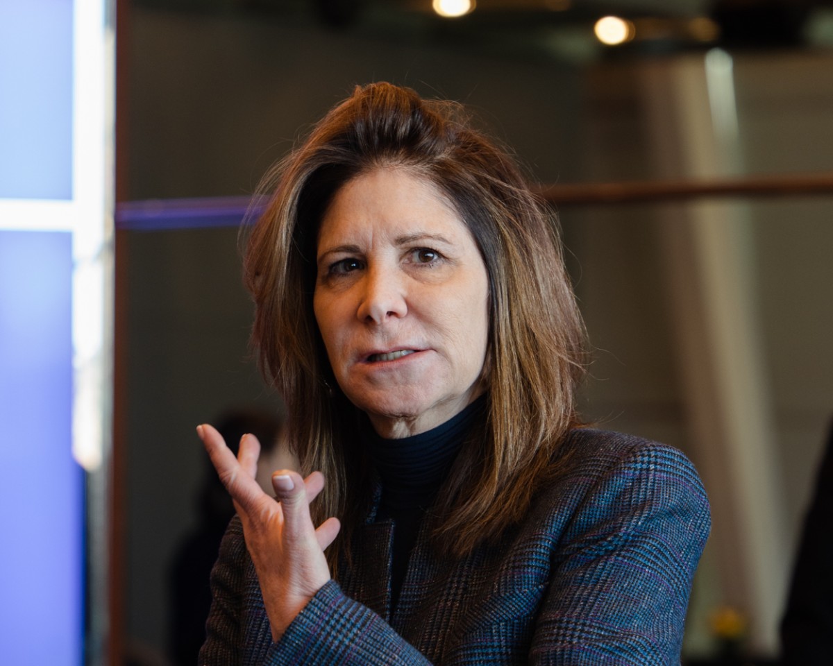 A woman stands with her left hand in the air. She is wearing a black blazer with grid patterns on the. She has short, brown hair.