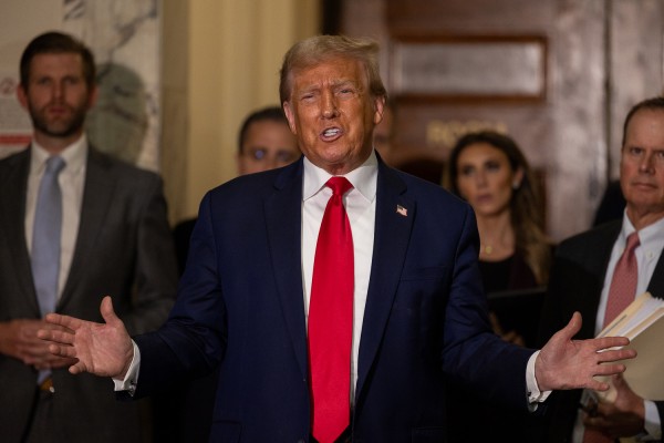 Donald Trump wearing a blue suit with a red tie, an American flag pin on his lapel and white undershirt with his arms outstretched. Behind him to the left is his son Eric Trump in a gray suit with a light blue tie, as well as his legal team all standing in front of a large brown door with golden words on them.
