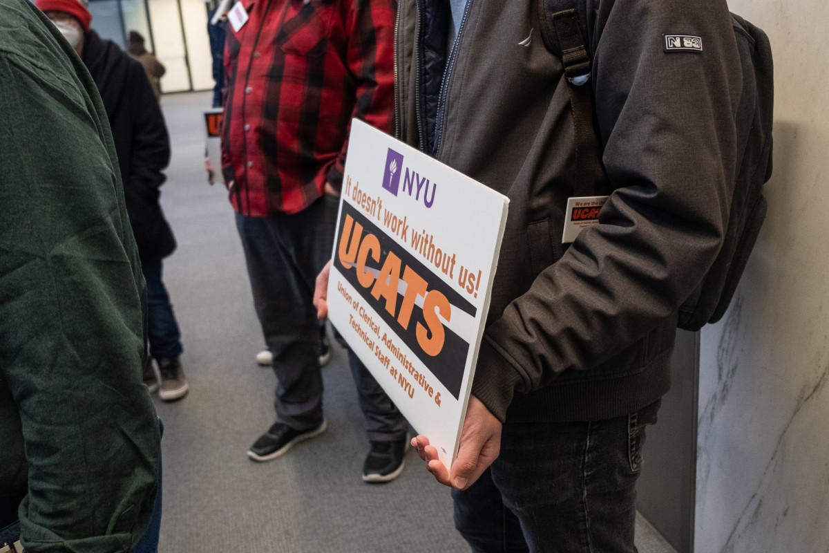A person wearing a gray jacket holds a sign reading “N.Y.U. It doesn’t work without us! U.C.A.T.S. Union of Clerical, Administrative and Technical Staff at N.Y.U.”