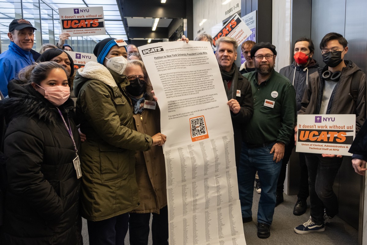 Two people standing in front of a crowd, hold up a large petition to N.Y.U. president Linda Mills, followed by a long list of names that signed it.
