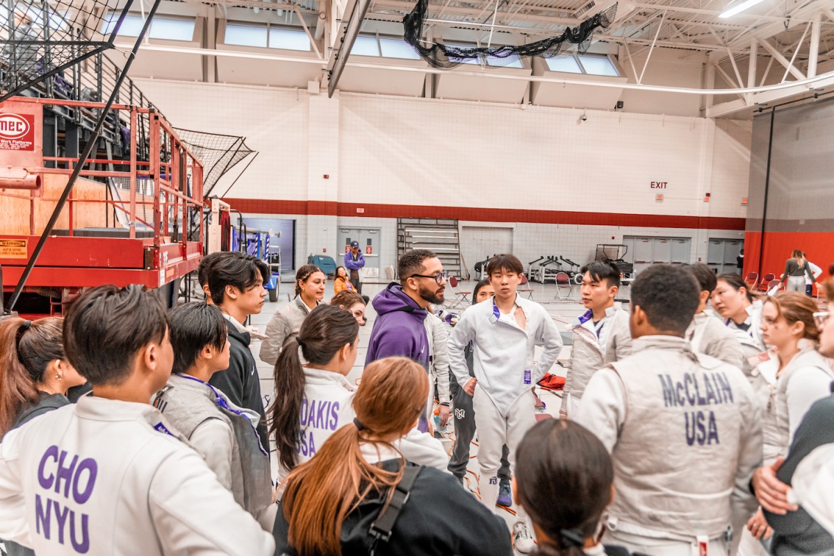 A group of men and women in white fencing suits and gray jackets are standing around a man in a purple sweatshirt with the Nike logo. They are all standing in the middle of a gym with a red line across the walls.