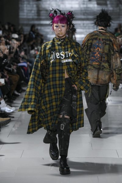 A model wears a black, yellow and green plaid jacket that says “Destroy Everything,” black pants, platform black shoes and a black choker.