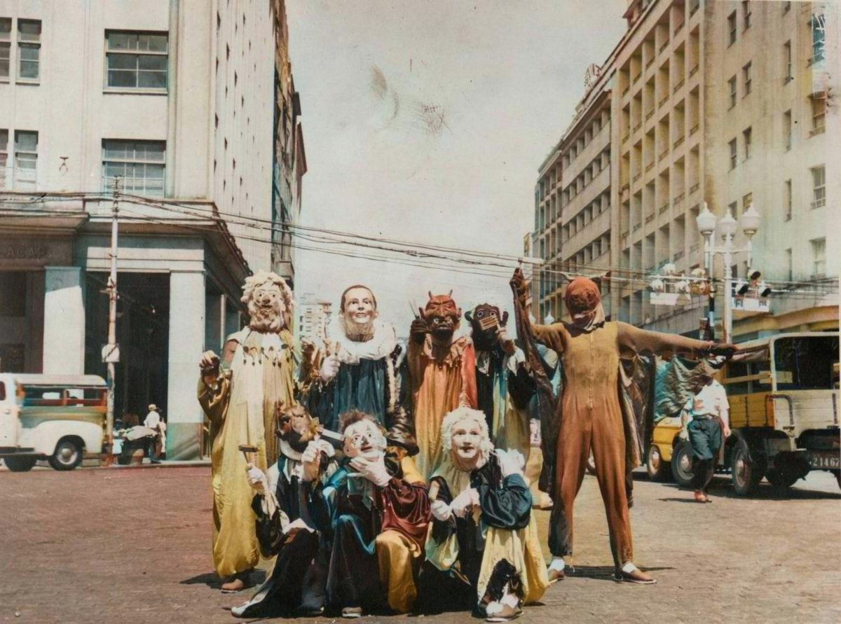 A faded, sepia-tinted film photograph with eight figures in medieval costumes. The figures stand in the middle of the road with vehicles and city buildings in the background.