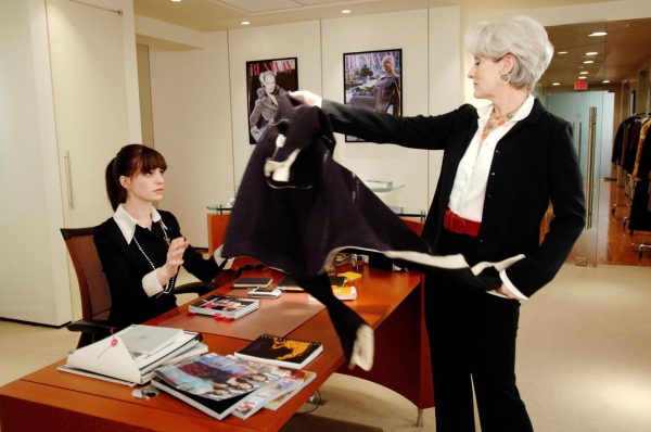 A woman stands in an office with fashion posters displayed on white closet doors, tossing her jacket to a woman sitting at a desk.