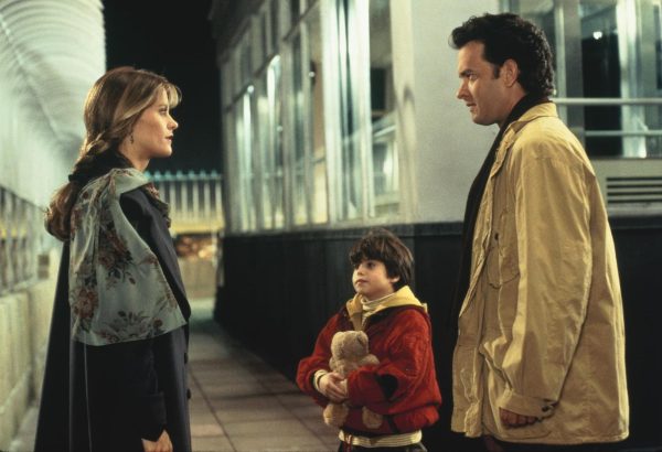 A woman wearing a dark blue jacket with a light blue scarf with floral print and a man wearing a beige jacket talking to each other at a train station. A little boy holding a teddy bear stands beside the man while looking at the woman.