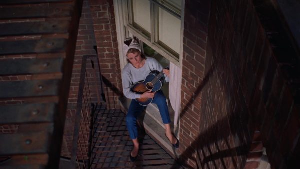 A bird’s-eye shot of a woman playing the guitar on her balcony. She is wearing blue jeans and sitting in her window.