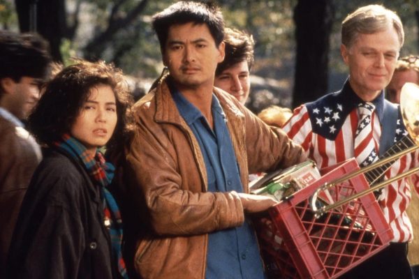 A man holding a red milk crate stands beside a woman wearing a scarf. They are both looking at some unseen subject in the distance. A man wearing an American flag shirt while holding a guitar stands beside them.