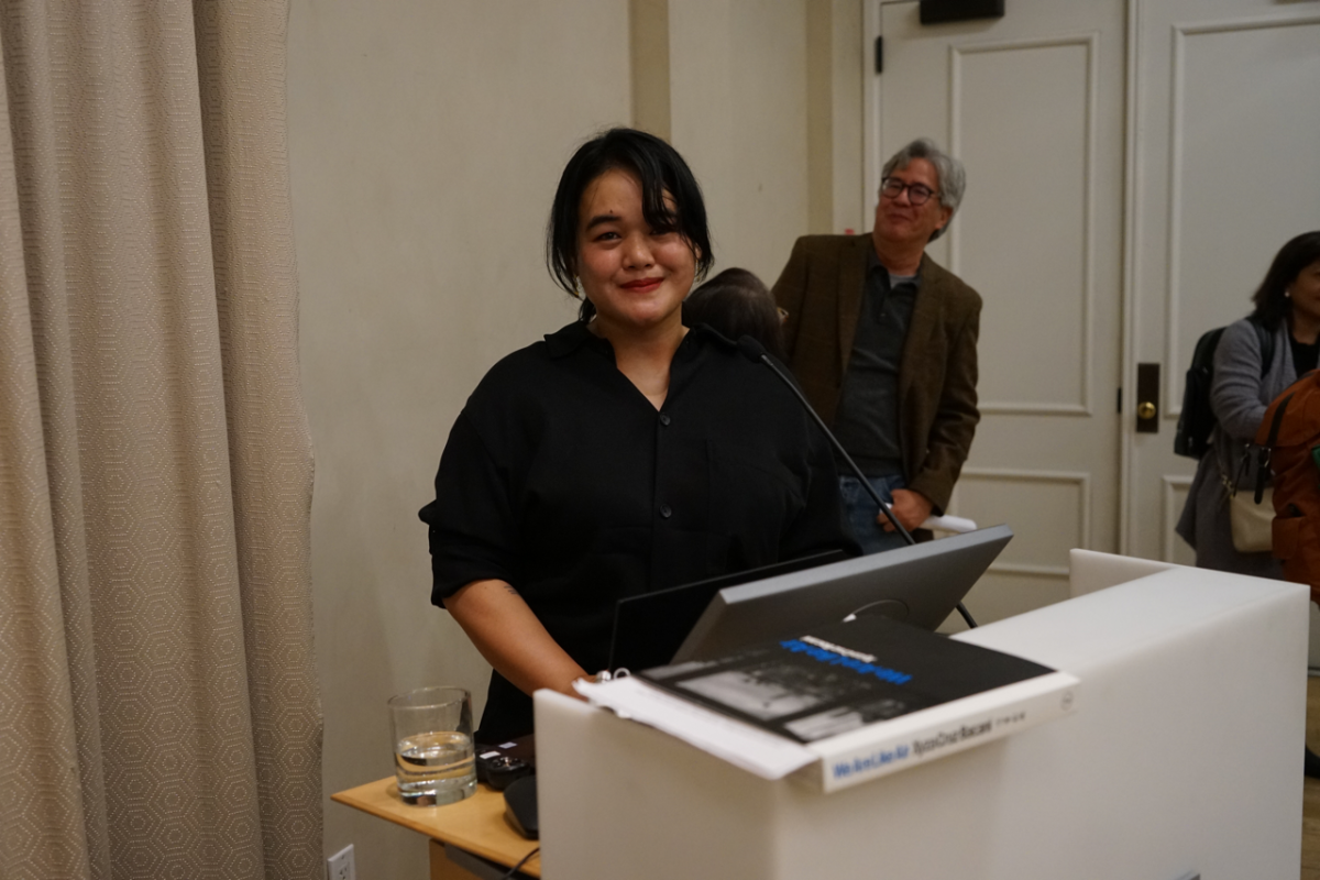 A woman with black hair wearing a black shirt stands behind a white podium in a lecture hall.