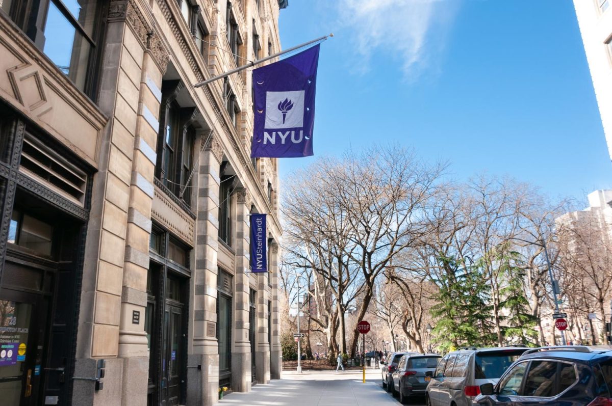 An N.Y.U. flag hangs above the Steinhardt School of Culture, Education and Human Development. Cars line the road to the right of the building and an entrance to Washington Square Park can be seen in the background.