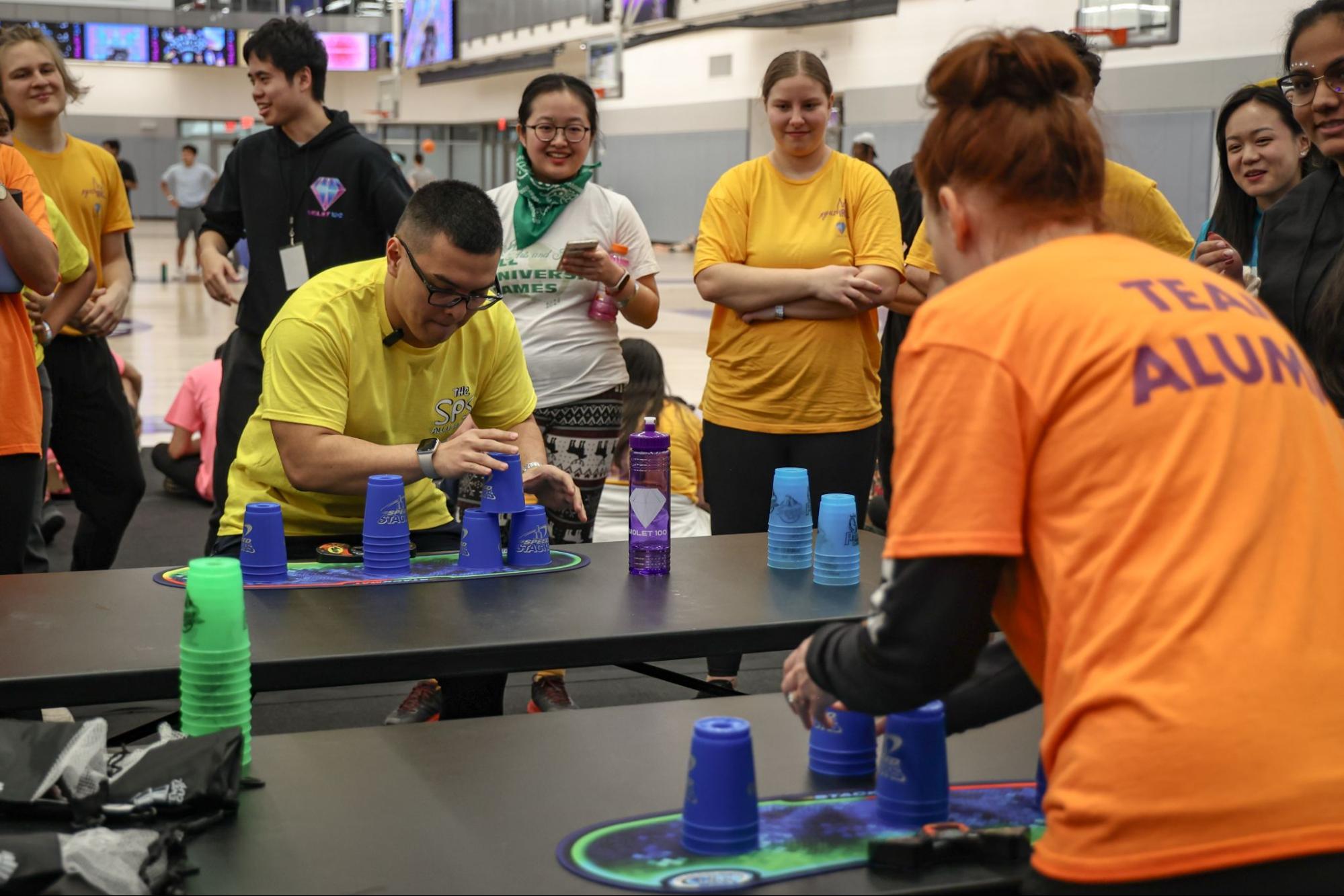 A man in yellow and a woman in orange stack cups across from each other while people stand around them watching.