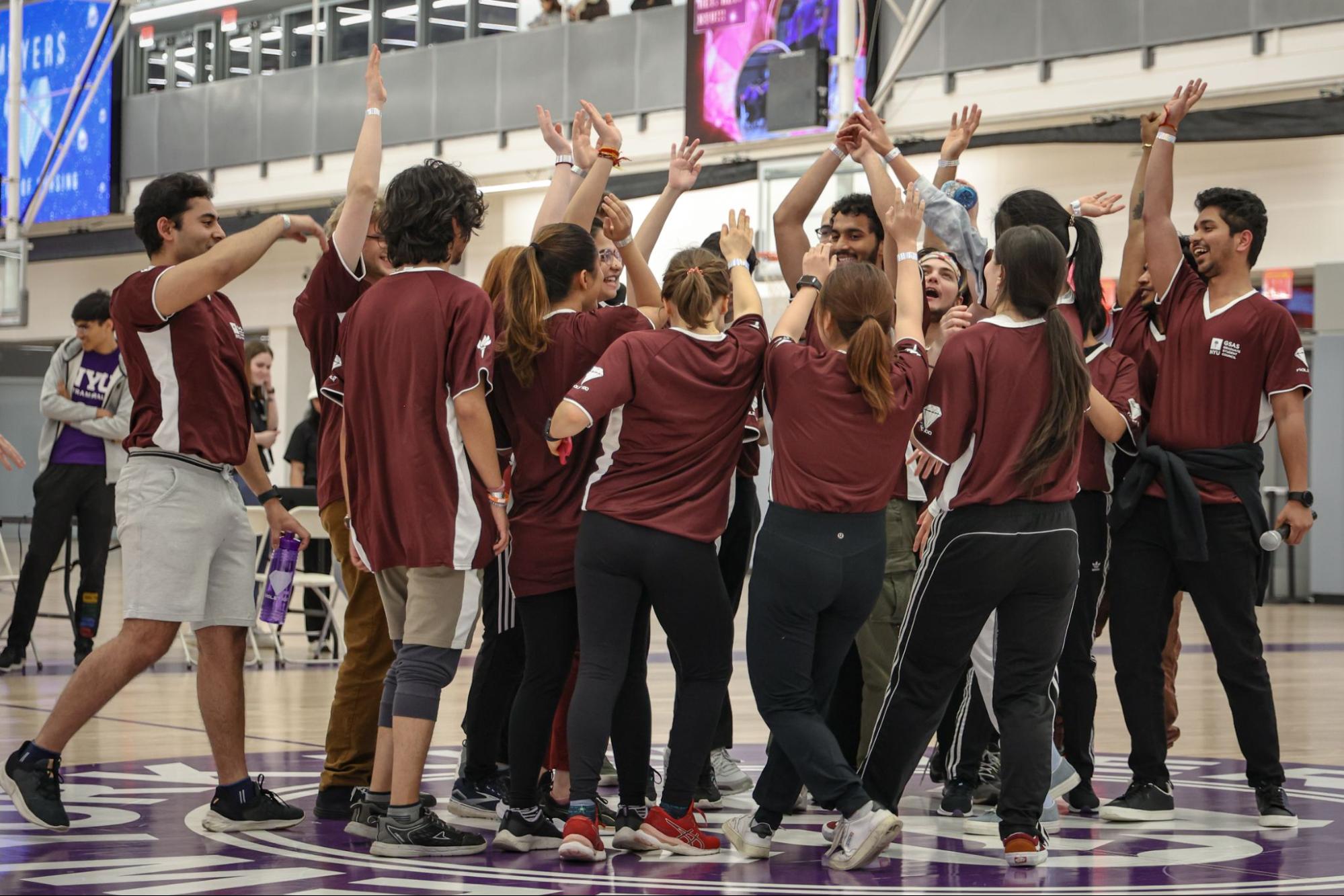 A team in maroon shirts stand in a circle with their hands raised. Their shirts say in white font “G.S.A.S.” along with the N.Y.U. torch logo.