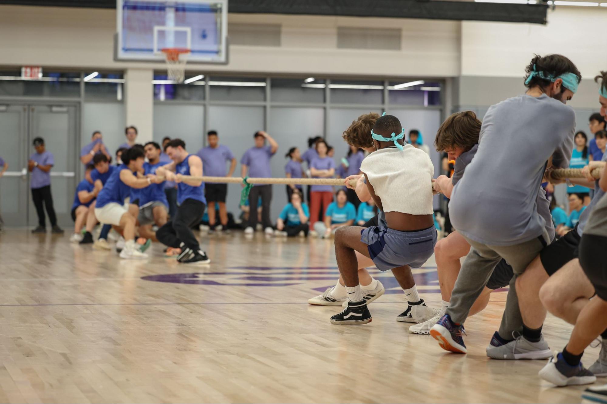A team in blue and a team in gray play tug-of-war on a basketball court. The teams pull from opposite sides of a beige rope with a green bandana tied in the middle.