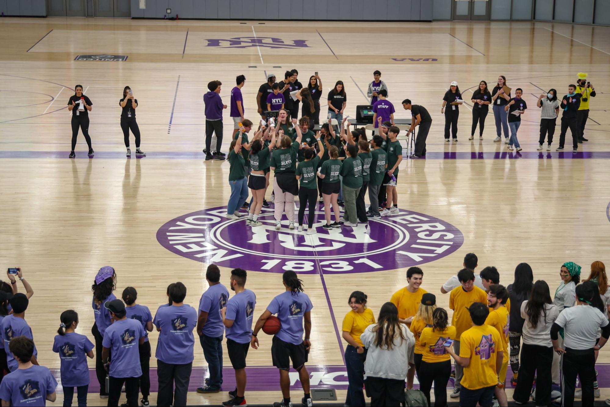 People stand in groups around a basketball court with the N.Y.U. logo. A group wearing matching green shirts stands in a circle at the center of the court.