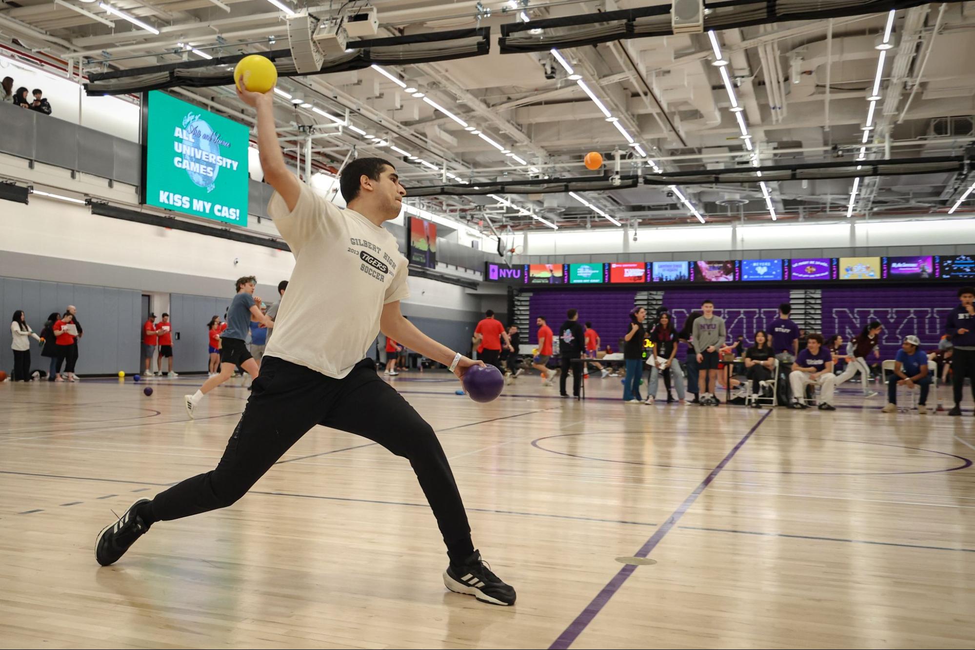 On a basketball court, a man winds up to throw a yellow ball while holding a purple ball in his other hand. In the background people spectate from the court's perimeter.