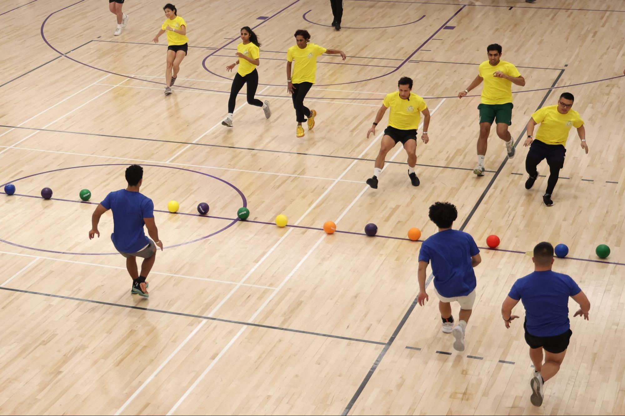 Two teams of people run towards a line of multi-colored balls at the center of a basketball court. One team wears purple and the other wears yellow.