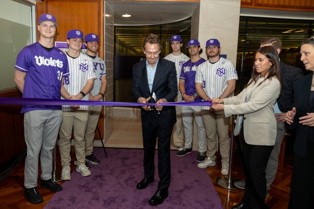 A man in a suit cutting purple ribbon while N.Y.U. baseball players stand behind him and watch.