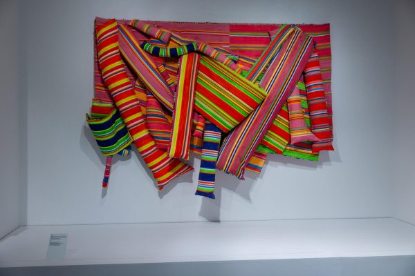 An art piece of overlapping, multicolored, striped fabric pieces hangs from a white wall.
