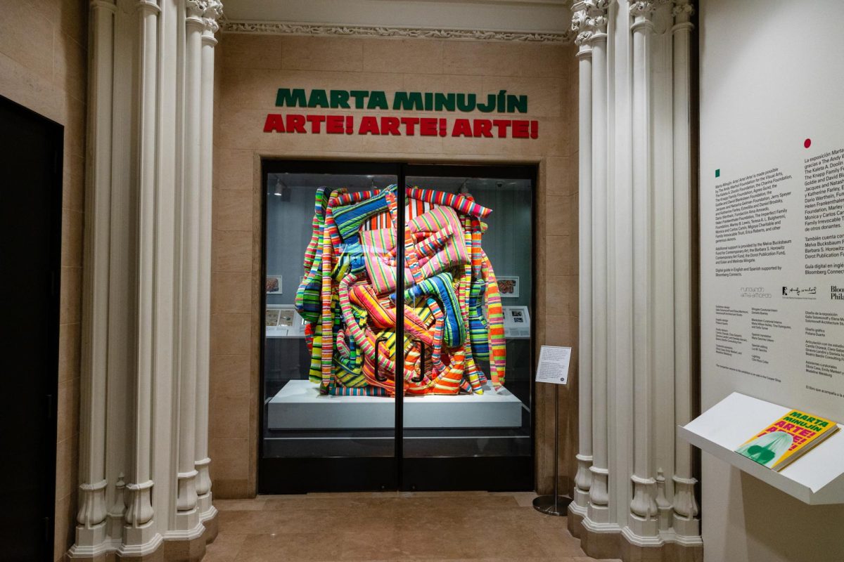 Museum display with headline “MARTA MINUJÍN: ARTE! ARTE! ARTE!” in green and red. Under the headline a glass display has a fabric sculpture of multicolored stripes.