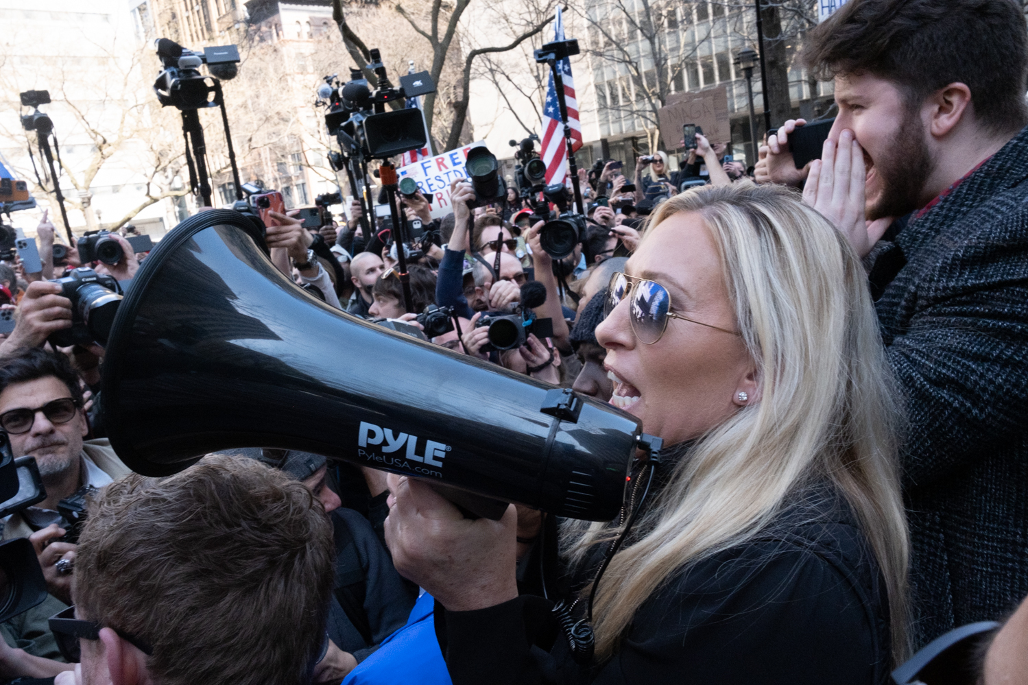 Representative Majorie Taylor Greene speaking to a crowd of protestors with a megaphone. There are many people pointing cameras towards her.