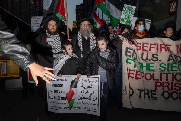Two children and two men hold a banner with Arabic and English written on it. Besides them a crowd carries a brown banner and Palestinian flags.