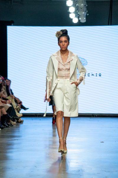 A model walking down a runway with a white blazer jacket with short white sleeves and straps on top of longer peach sleeves, on top of a silky, peach button-up shirt.