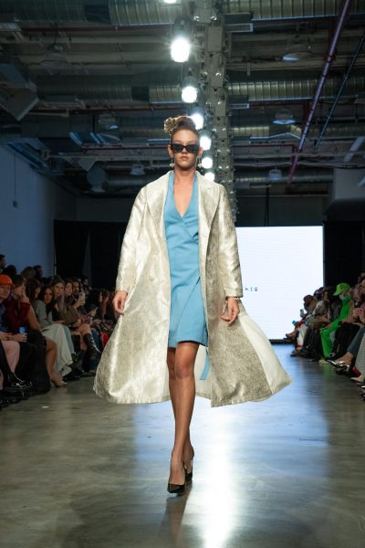 A model walking down a runway with a sky-blue dress and a shiny, oversized silver jacket.