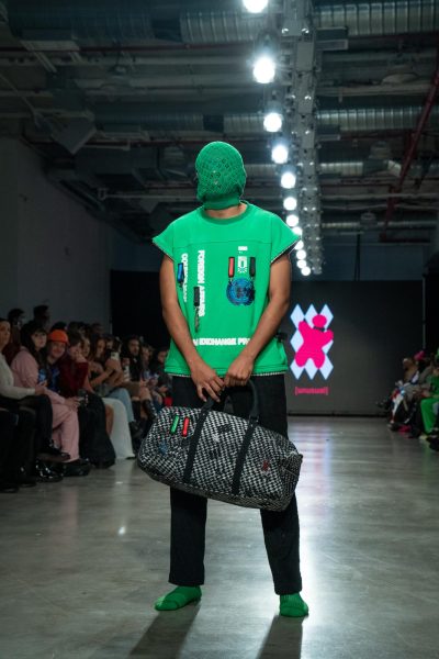 A model is wearing green socks, black pants, a green shirt and a green face mask that covers their whole face. The model is also holding a checkered bag.