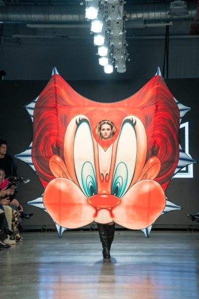 A model walking down a runway and wearing an image of a cartoon face that has red hair.