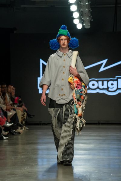 A model walking down a runway wearing a fuzzy blue hat and a gray jacket with a second collar at the bottom of the shirt. The model carries a multi-colored string bag over his shoulder.
