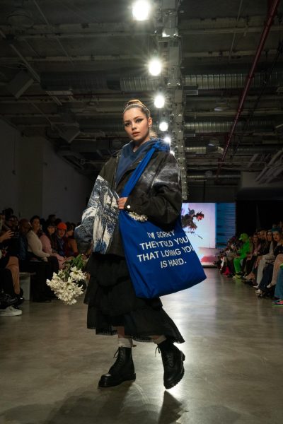 A model wears a blue hoodie and a black zip-up. The model also wears a long ruffled black skirt and has a blue tote bag that says “I’M SORRY THEY TOLD YOU THAT LOVING IS HARD.”