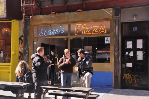 Exterior shot of a pizza shop with multiple people and picnic tables in front of it. Above the entrance is a blue and yellow neon sign that says “Scarr’s Pizza.”