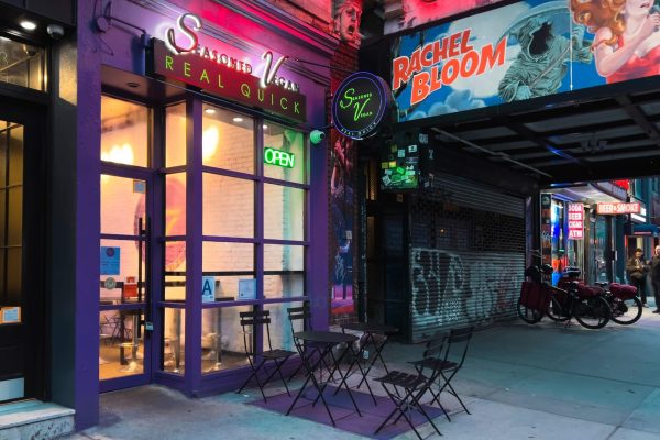 A purple storefront of a restaurant with the name “Seasoned Vegan REAL QUICK” placed above the entrance. Black chairs and tables are placed in front of it.