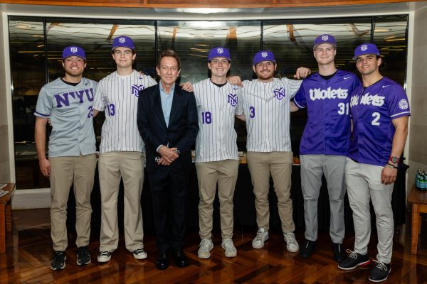 A man in a suit stands with six men in N.Y.U. baseball jerseys and purple N.Y.U. hats.