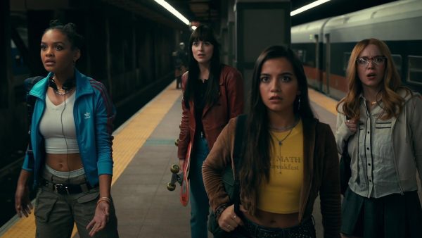 Four women looking scared on a subway platform. One of them is holding a skateboard.