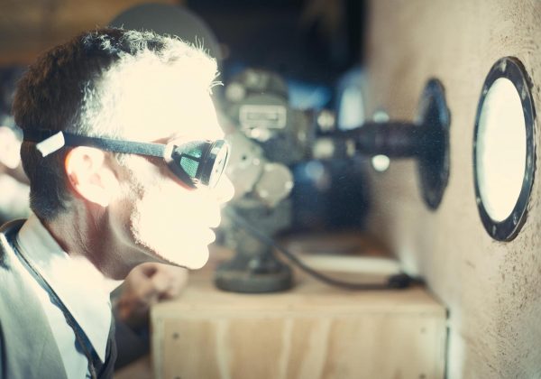 A man wearing protective goggles looks through a small circular window that is emitting bright light.
