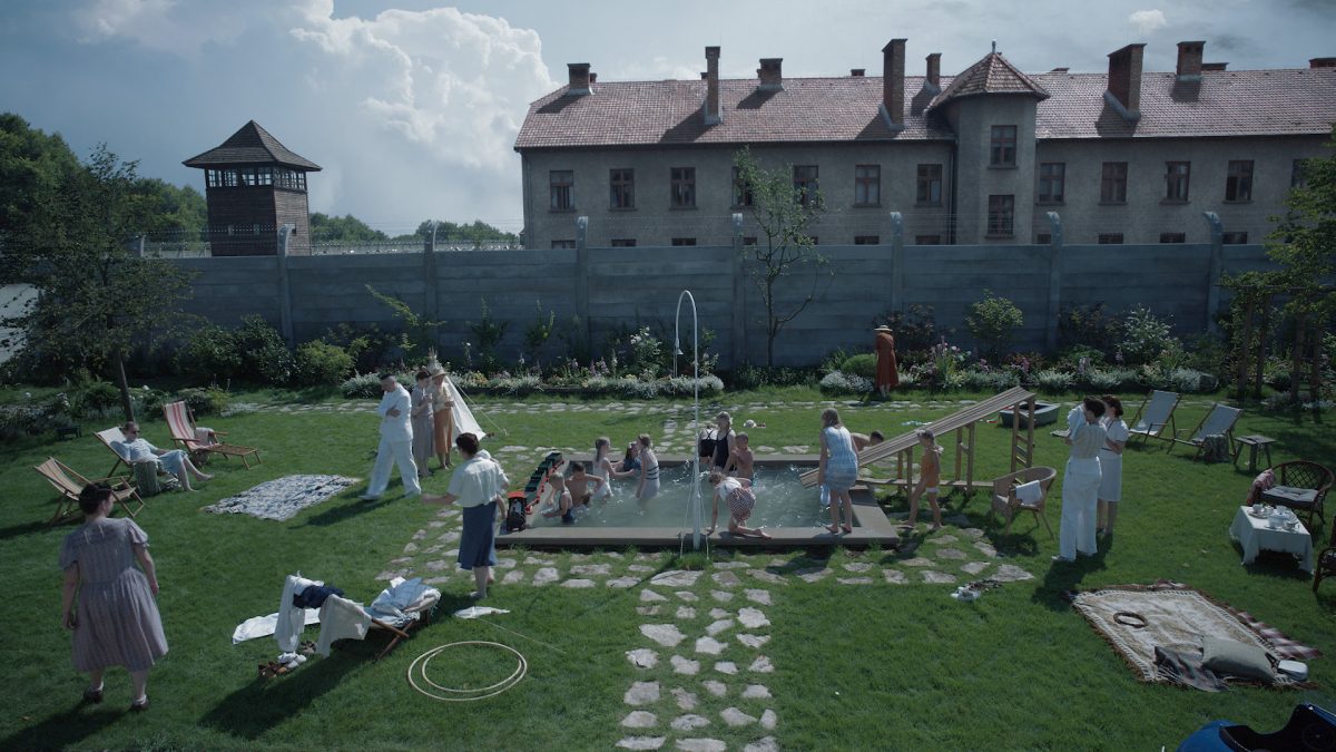 A garden with green grass, chairs and stone paths, surrounds a shallow pool that children are playing in while adults watch. Behind the garden, there is a wall with barbed wire, guard towers and a large red-shingle roofed building.